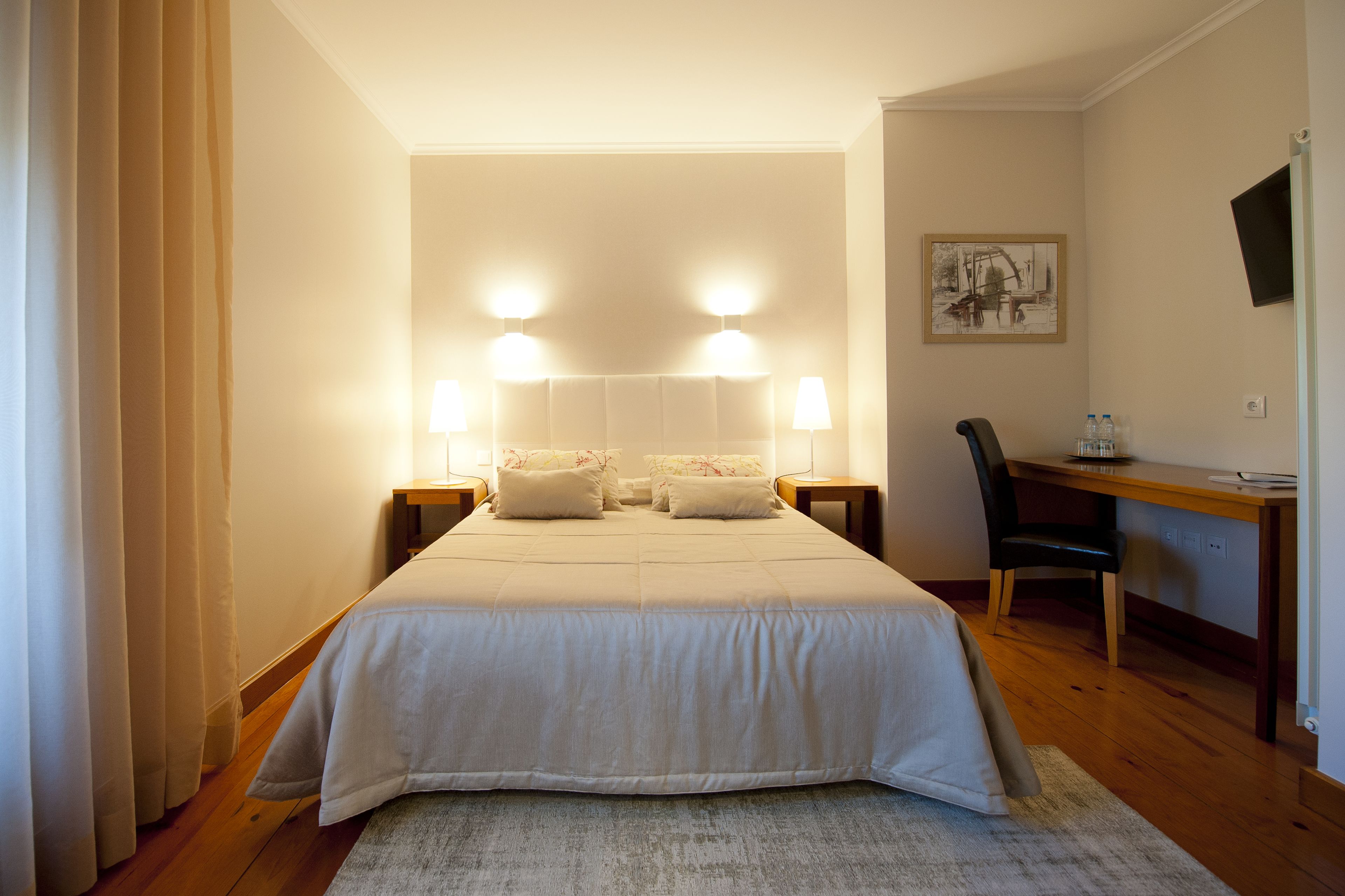 A hotel room with a large bed, two bedside lamps, a desk with a chair, and a framed picture on the wall.