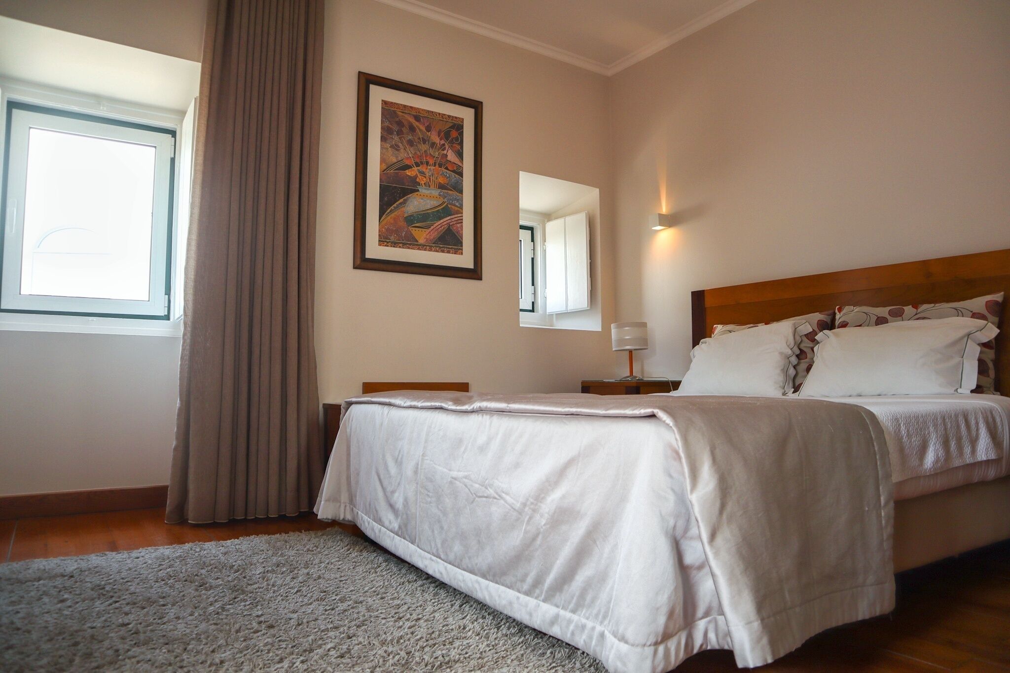 A bedroom with a bed, a nightstand, a bedside table, a mirror, a window with curtains, and a painting on the wall.
