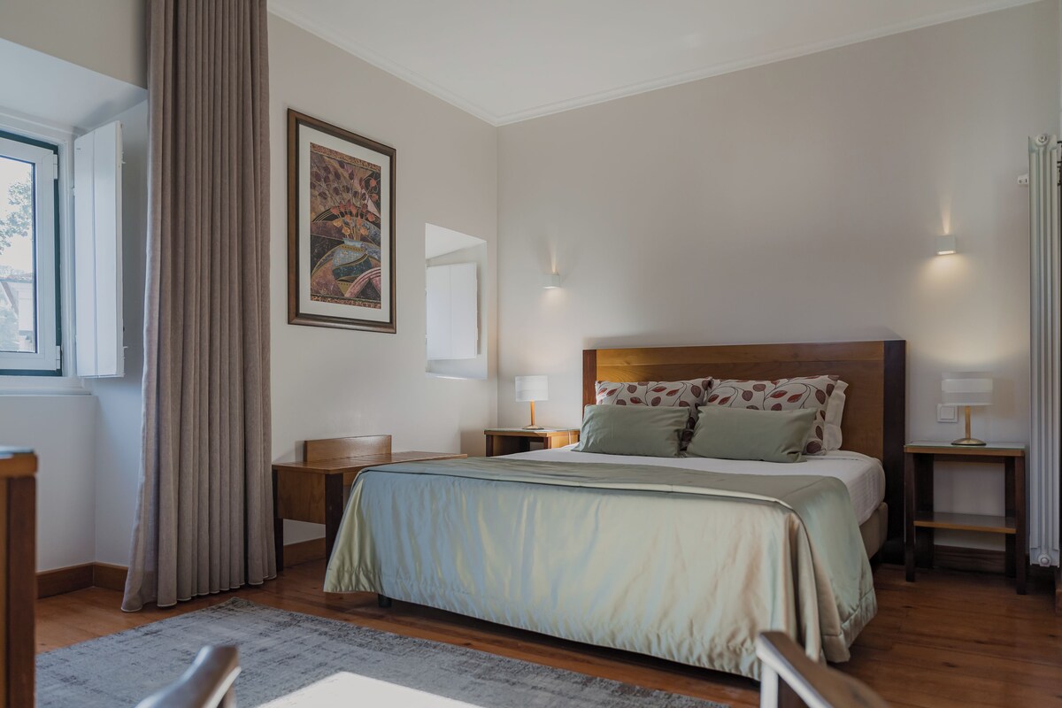 Un dormitorio con cama, mesitas de noche, lámpara, ventana con cortinas e imagen enmarcada en la pared.