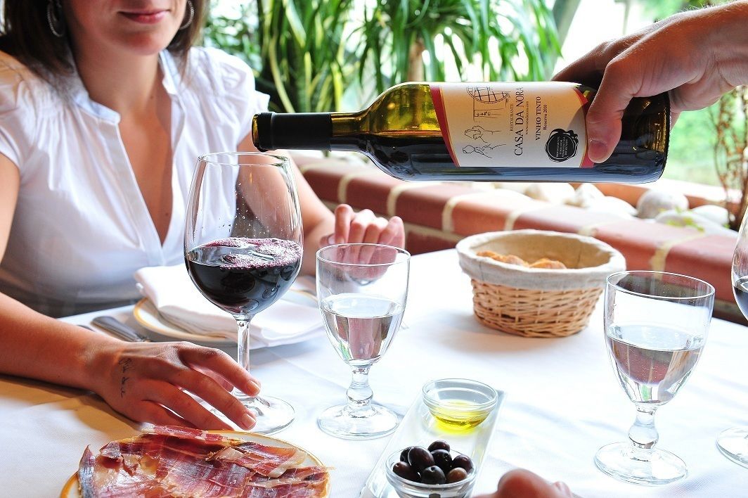 A dining table with a bottle of wine being poured into a glass, a basket of bread, olives, and a plate of cured meat.