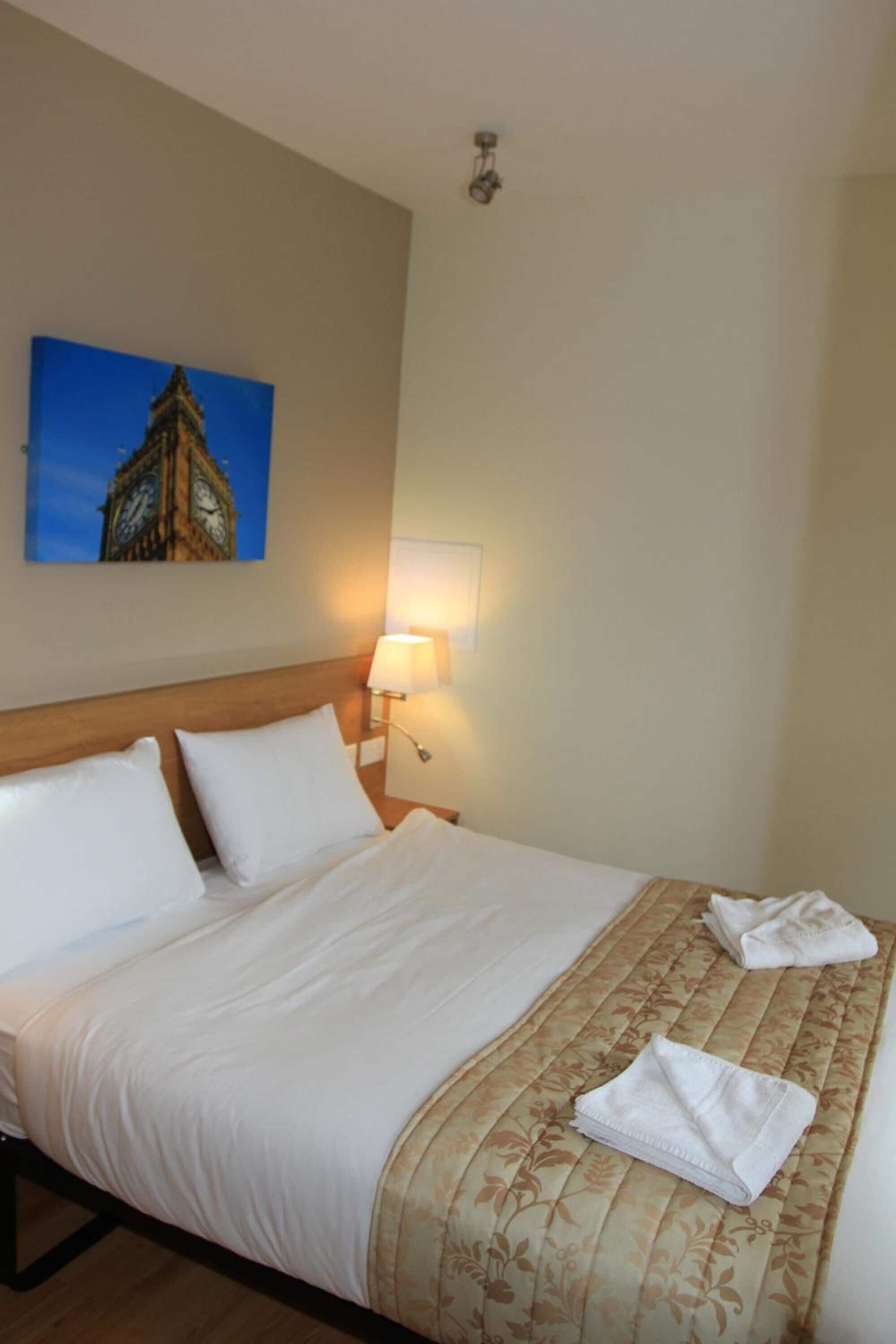 A hotel room with a bed, a nightstand with a lamp, and a painting of a clock tower on the wall.