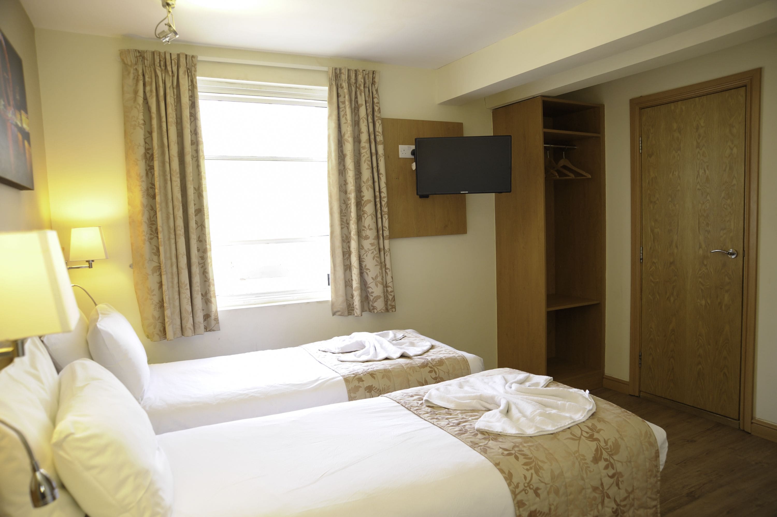 Habitación de hotel con dos camas, un televisor montado en la pared, un armario y una ventana con cortinas.