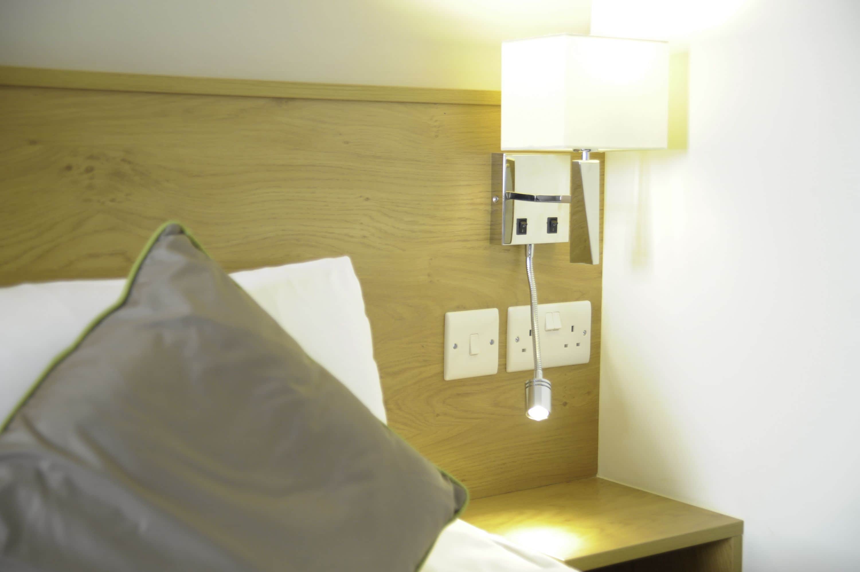 Een hotelkamer met een houten hoofdeinde, een bedlamp en een stopcontact.