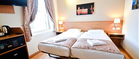 Quarto de hotel com duas camas, televisão, escrivaninha com computador e janela com cortinas.