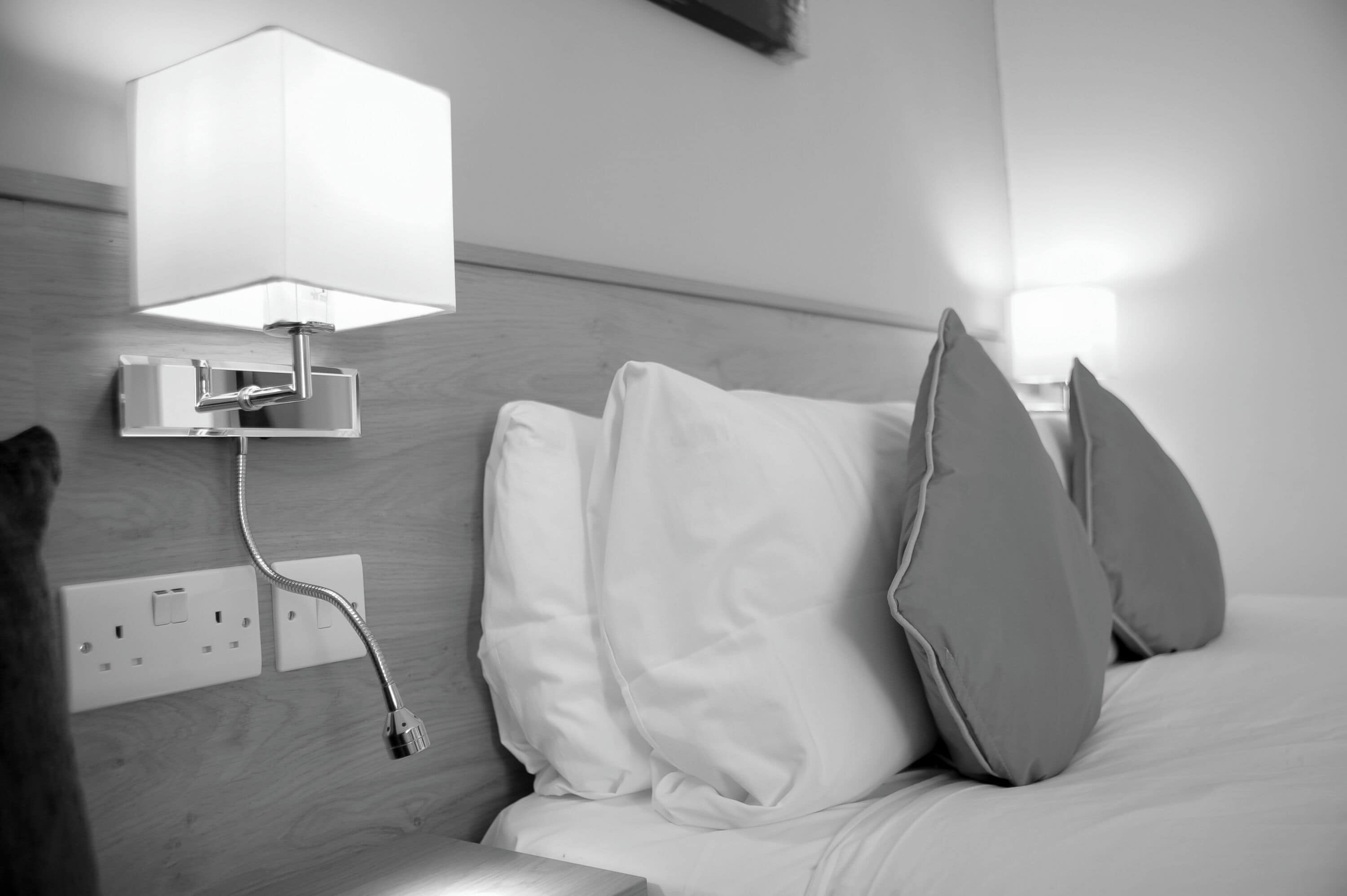 A hotel room with a bed, pillows, a nightstand, a lamp, and a power outlet.