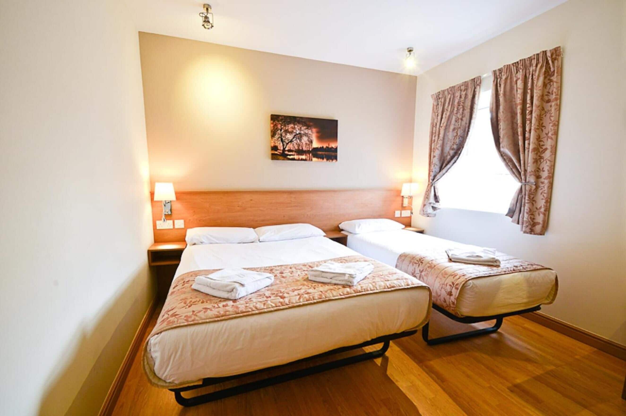 A double room with two beds, a wooden headboard, bedside lamps, a painting on the wall, and a window with curtains.
