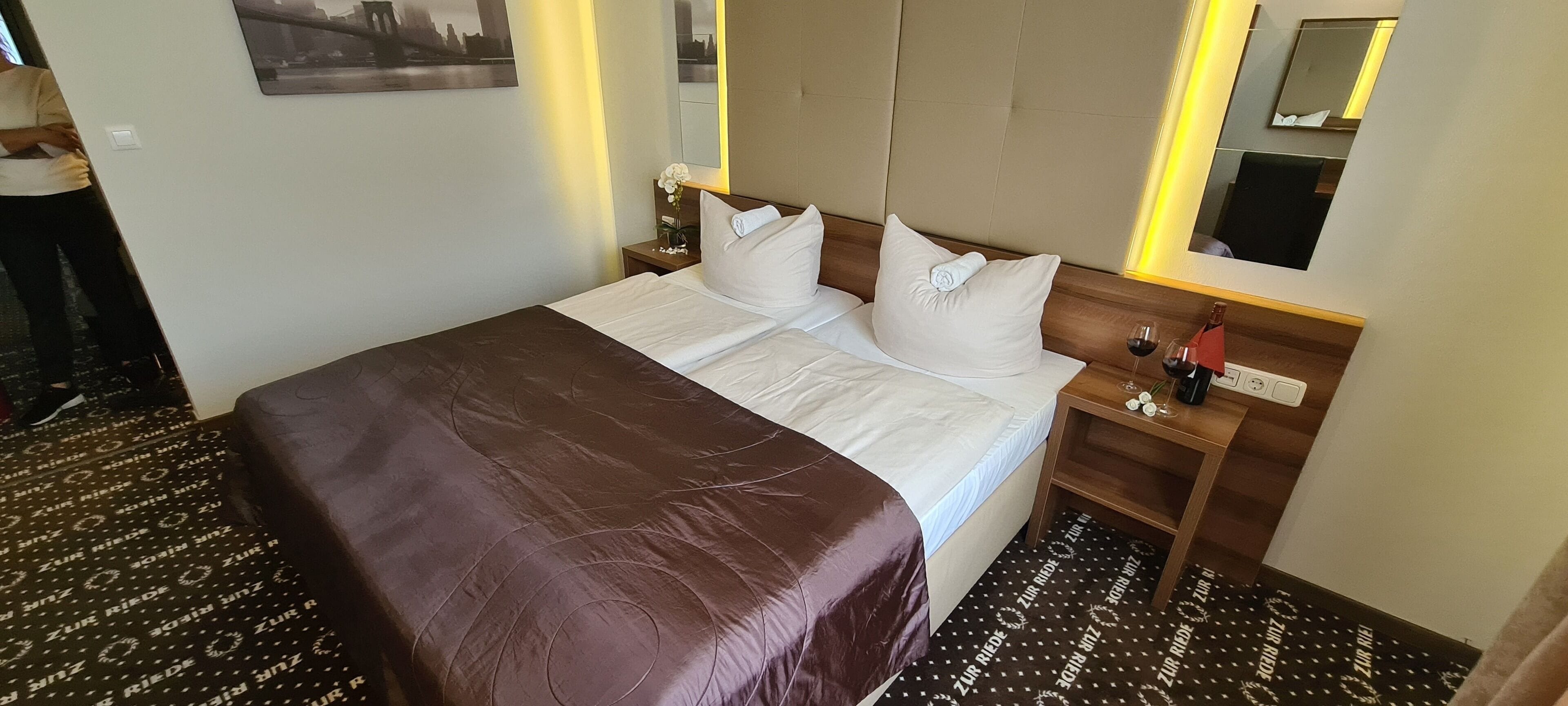 Une chambre d’hôtel avec un grand lit, une table de chevet avec une lampe, un miroir et une petite table avec un verre à vin.