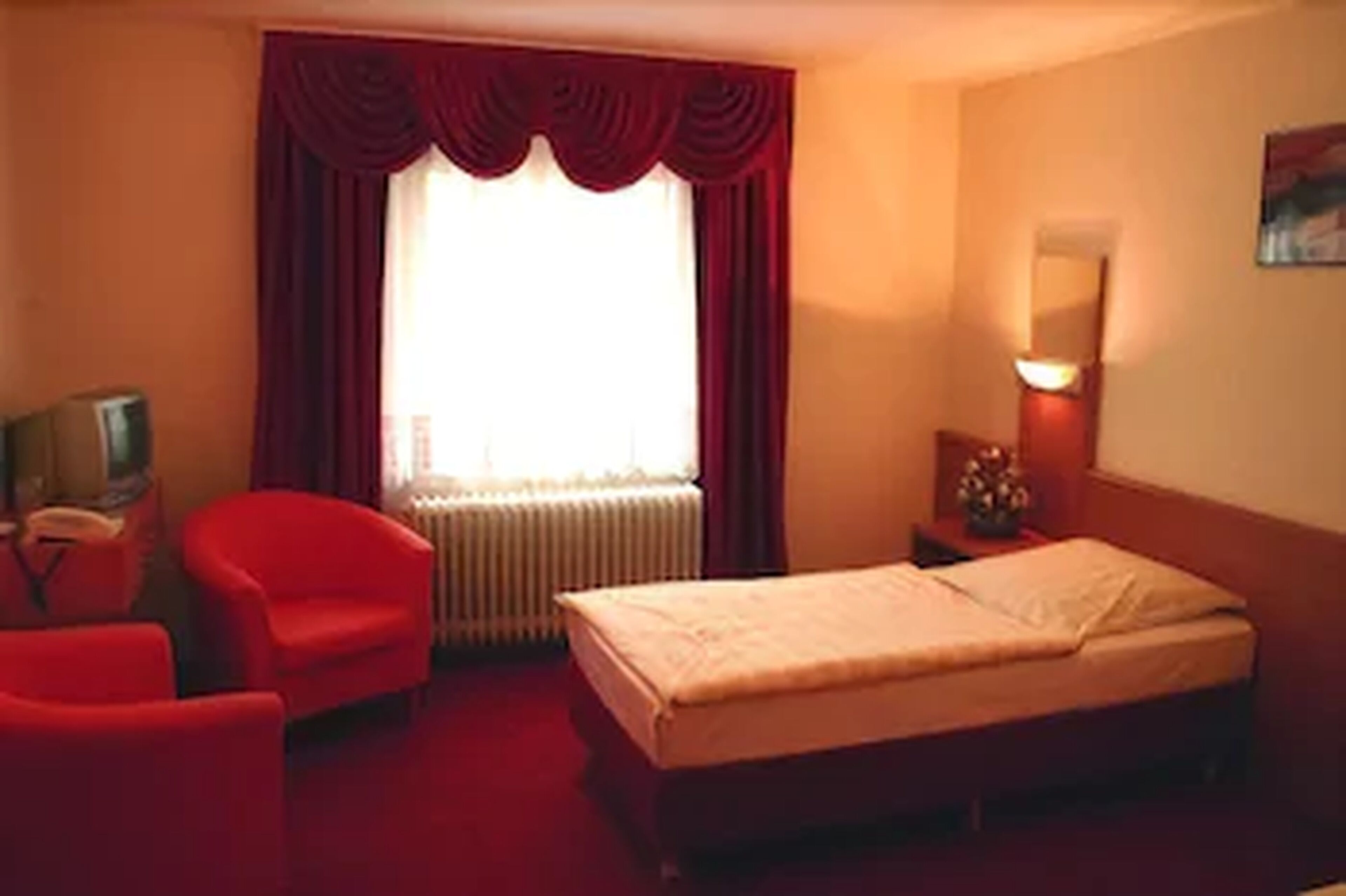 Habitación de hotel con cama, un sillón rojo, una mesita con lámpara y un radiador.