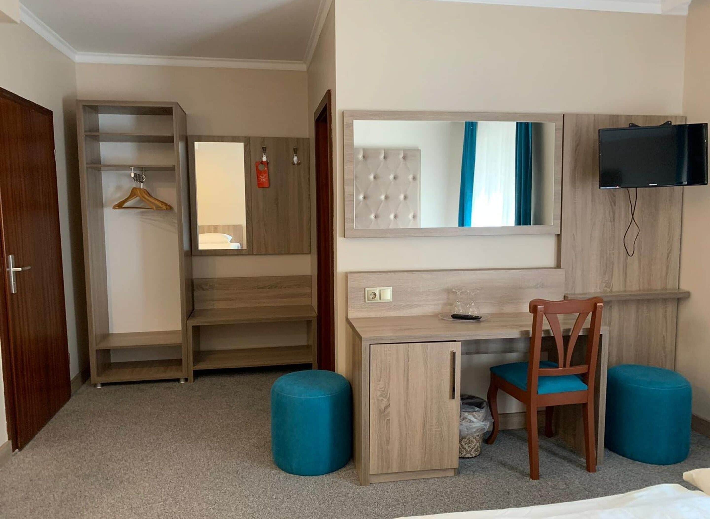 A hotel room with a wooden desk, a chair, a TV mounted on the wall, and a wardrobe with a mirror.