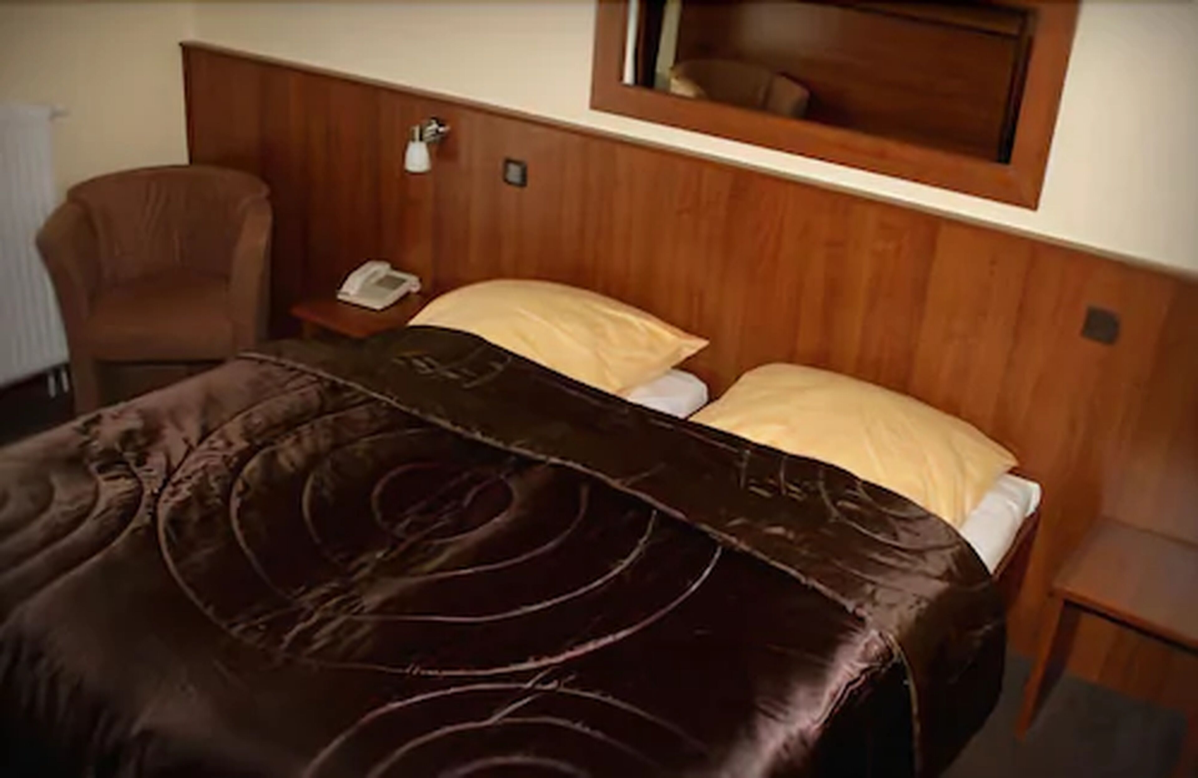 A hotel room with a wooden headboard, a brown leather bedspread, a bedside table with a telephone, and a chair.