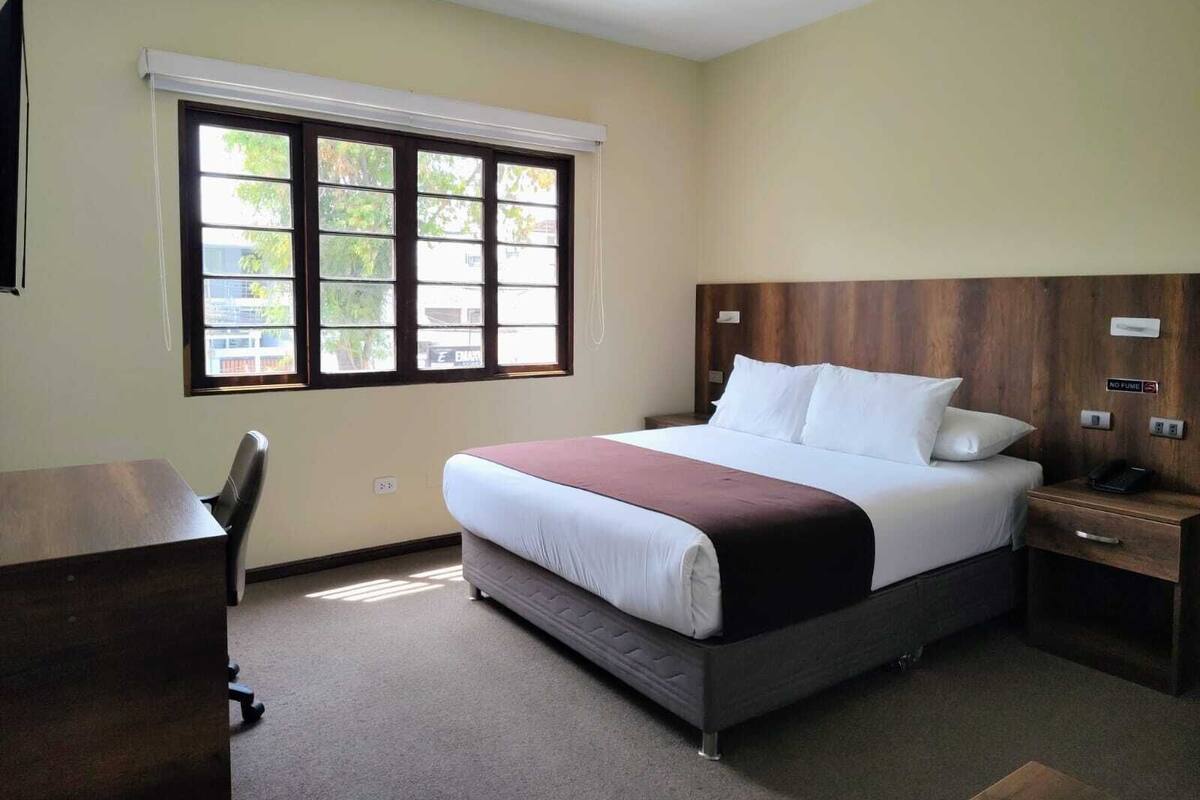 A hotel room with a bed, a desk, a chair, a nightstand, and a window with blinds.