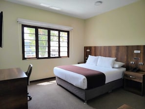 A hotel room with a bed, a desk, a chair, a nightstand, and a window with blinds.