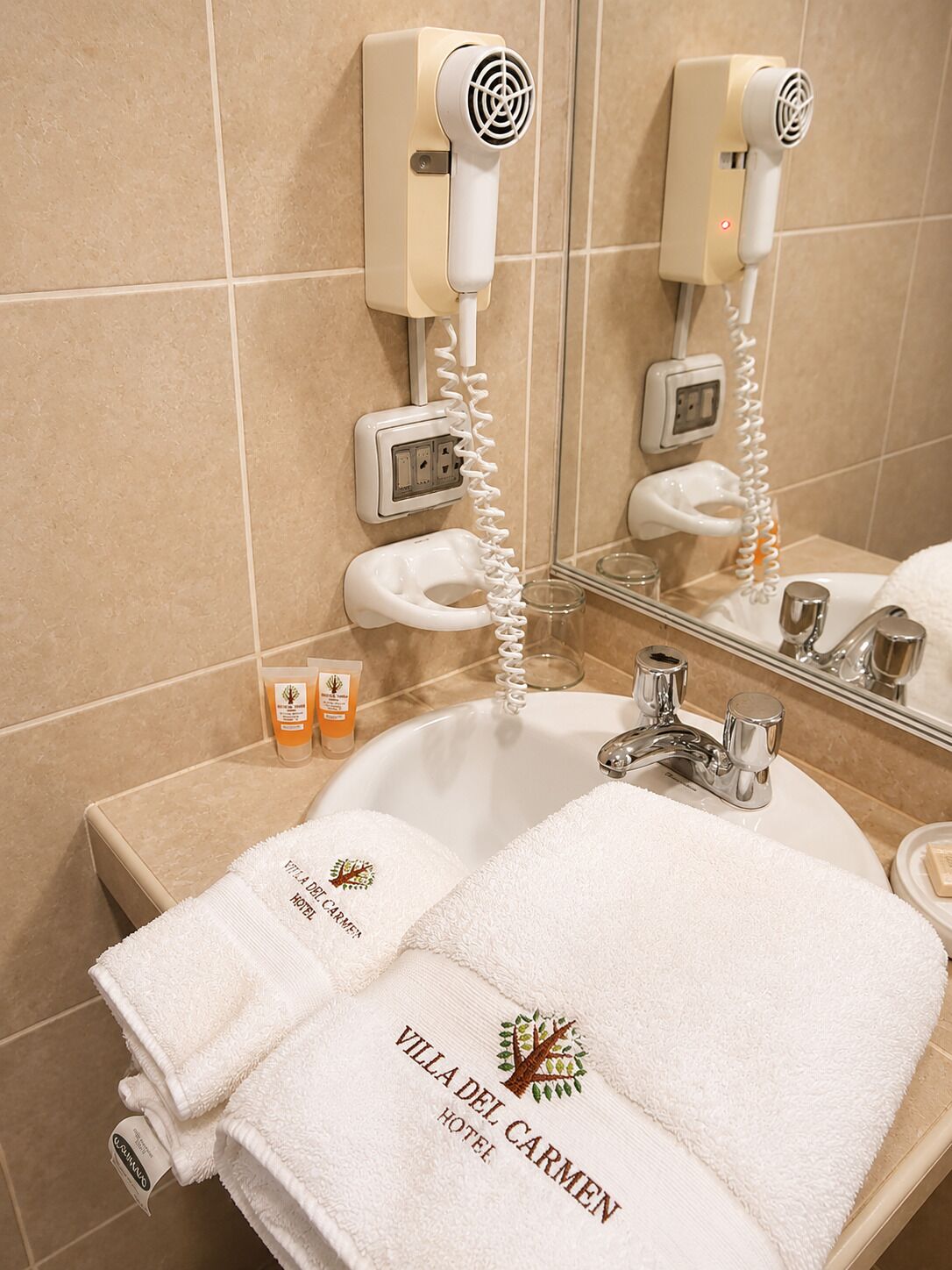 A bathroom with a sink, two hair dryers, a mirror, and two towels with 'Villa del Carmen Hotel' branding.