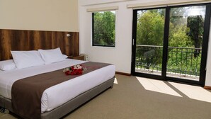 A hotel room with a large bed, a wooden headboard, a window with a view of greenery, and a balcony with a metal railing. - Hotel Villa del Carmen (Arequipa)