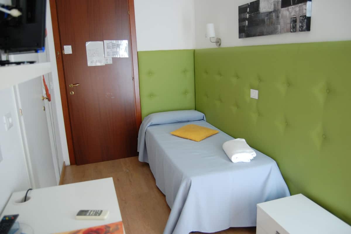 A small, single-bed room with a green headboard, a wooden door, and a white desk with a remote control.