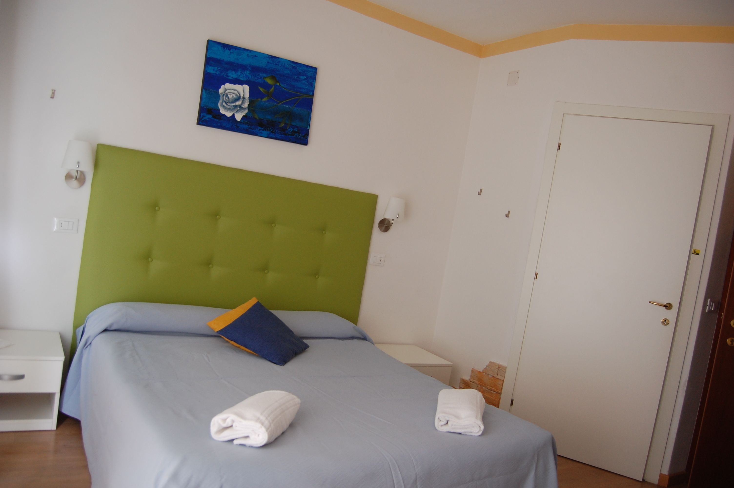Un dormitorio con un cabecero tapizado en verde, una cama individual con ropa de cama azul y blanca, dos mesitas de noche blancas, una puerta blanca y un cuadro azul en la pared.
