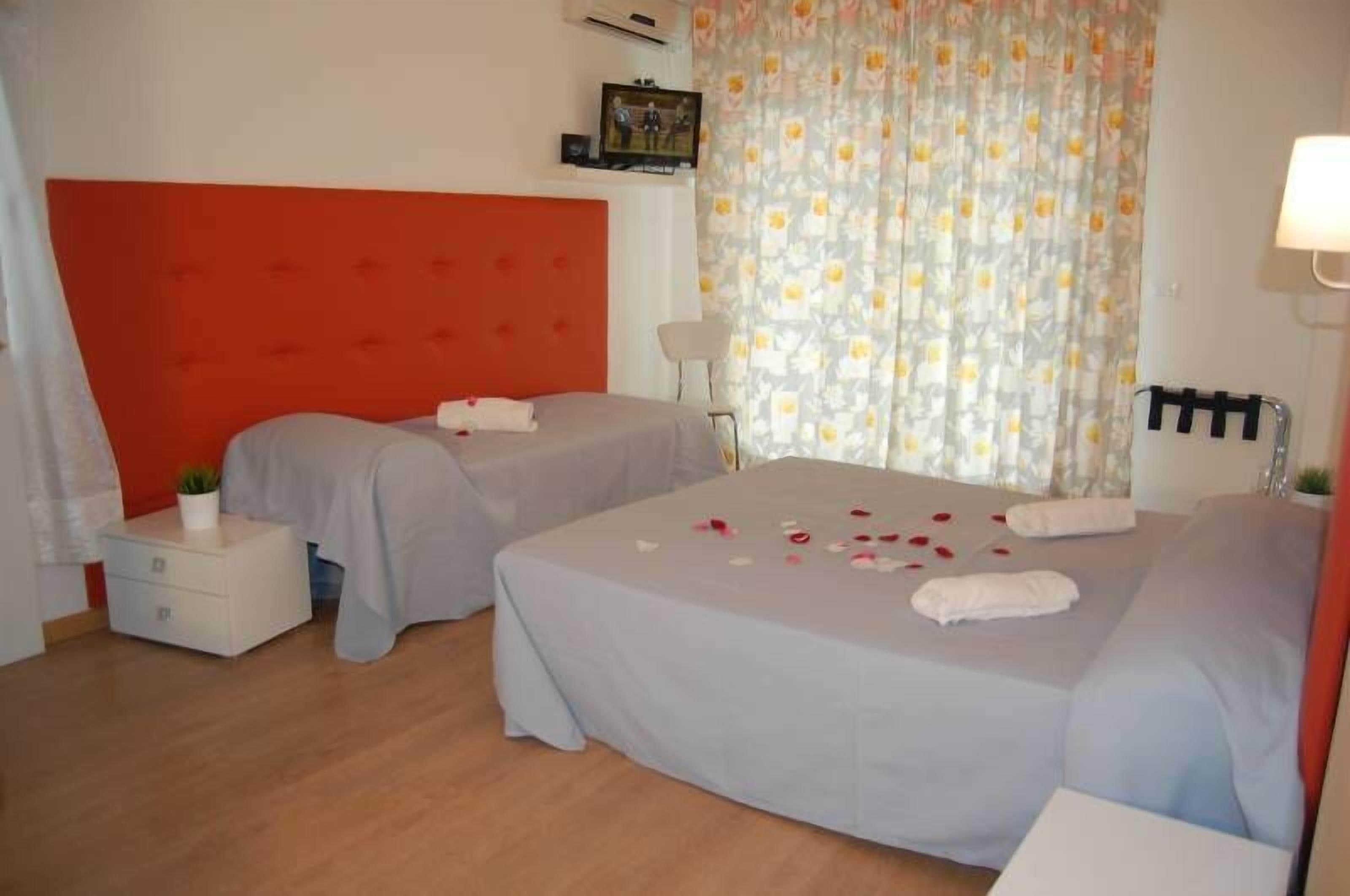 Een hotelkamer met twee bedden, een rode hoofdbord, een klein nachtkastje met een plant, en een raam met bloemen gordijnen.
