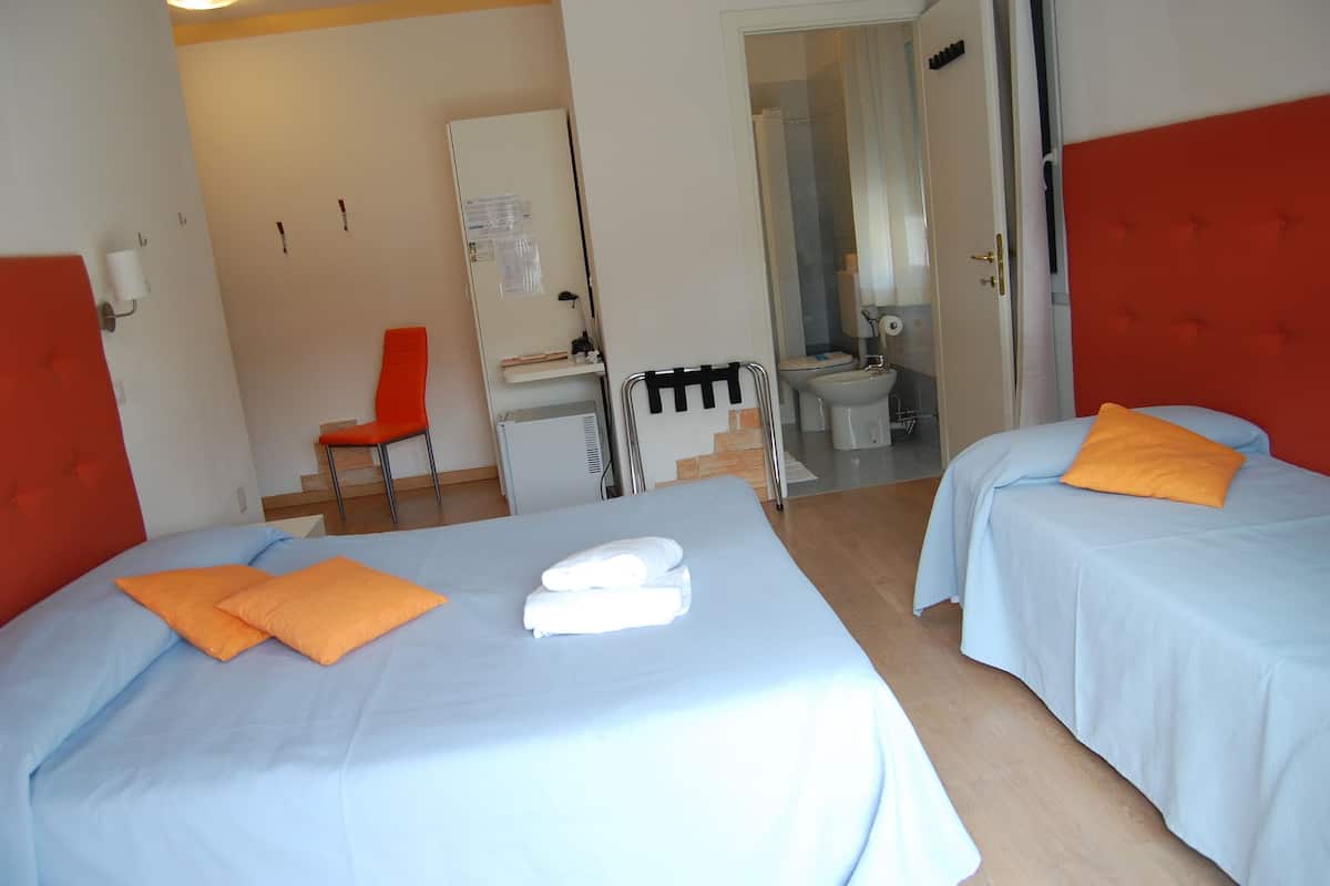 A hotel room with two beds, each with orange pillows and white towels. There is a desk with a chair, a toilet, and a shower visible through an open door.