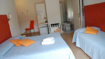 Habitación de hotel con dos camas, cada una con almohadas naranjas y toallas blancas. Se ve un escritorio con una silla, un inodoro y una ducha a través de una puerta abierta.