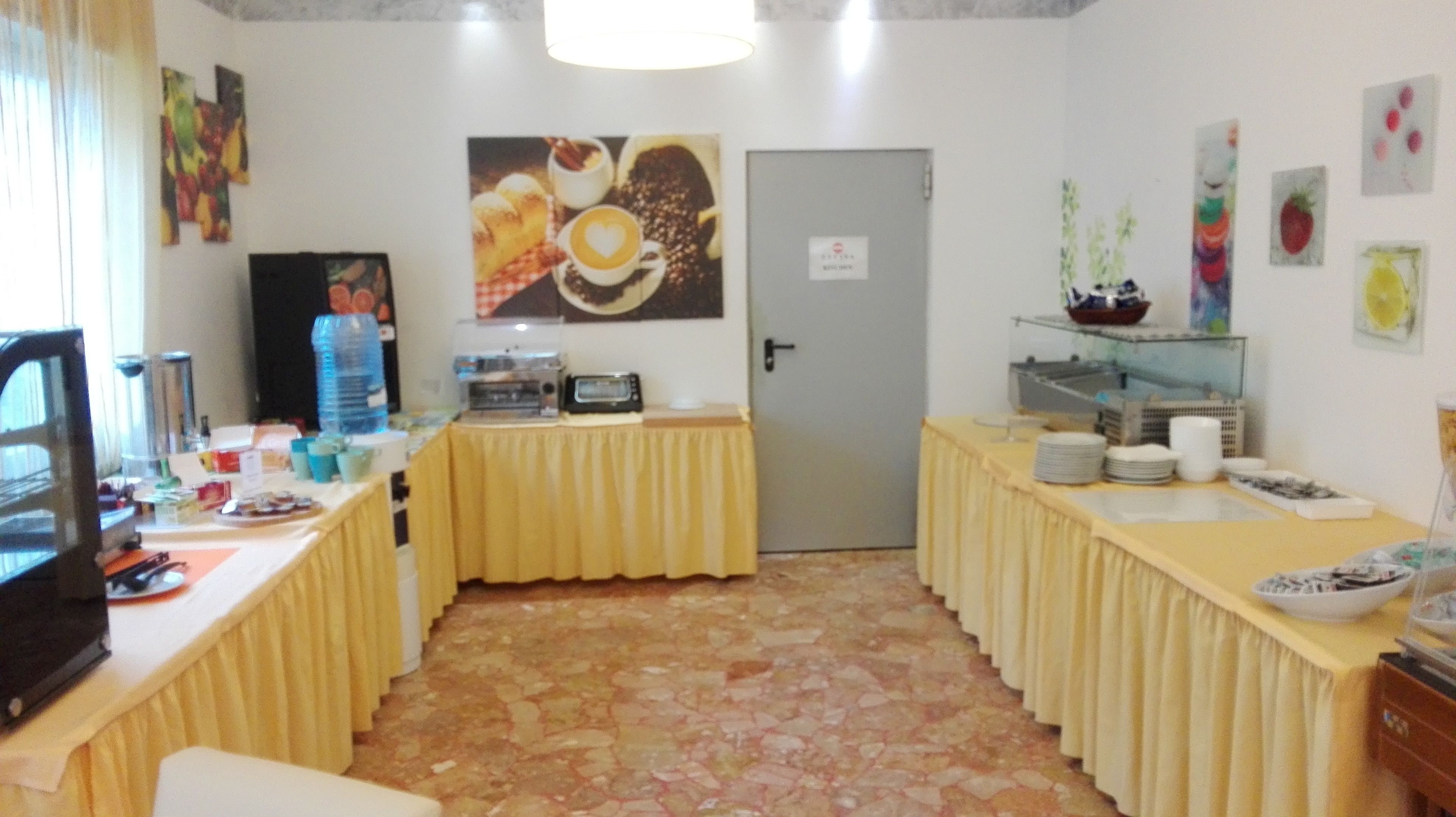Ein Zimmer mit Fliesenboden, einer Tür, einem Tisch mit gelber Tischdecke und verschiedenen Gegenständen darauf, darunter eine Kaffeemaschine, Tassen und eine Flasche.
