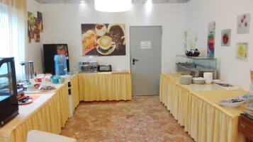 Ein Zimmer mit Fliesenboden, einer Tür, einem Tisch mit gelber Tischdecke und verschiedenen Gegenständen darauf, darunter eine Kaffeemaschine, Tassen und eine Flasche.
