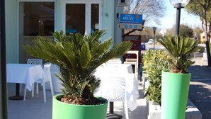 A hotel exterior with a bar sign, outdoor seating with white tables and chairs, and two potted plants. - Hotel Irene (Lignano Sabbiadoro)