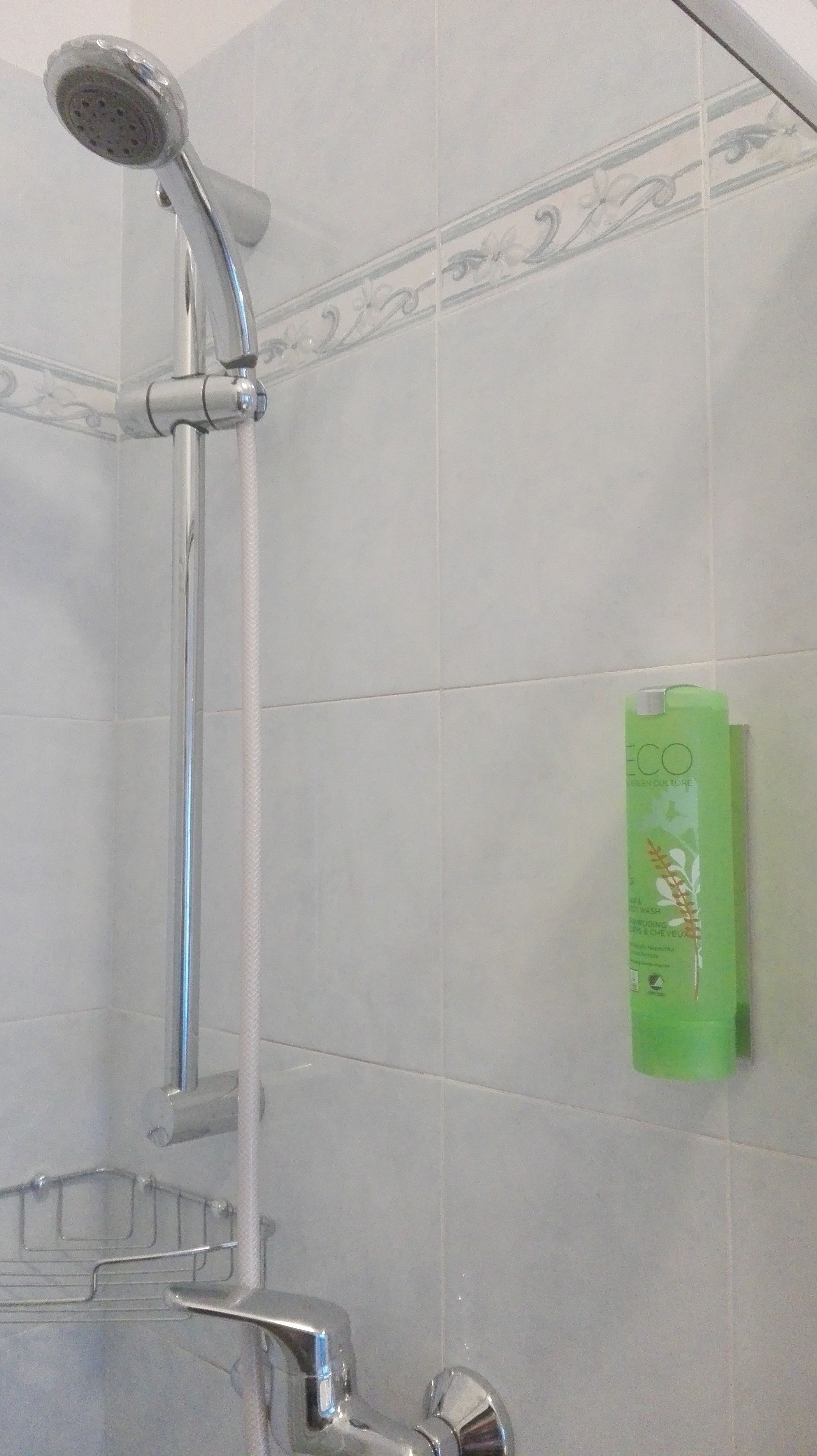 Een douche met een handdouche en een kraan op de muur. Een groene shampoo fles is zichtbaar aan de wand.