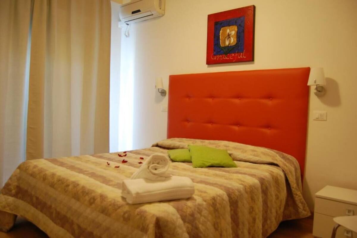 A hotel room with a large orange headboard, a bed with a striped bedspread, two green pillows, and a rolled white towel.