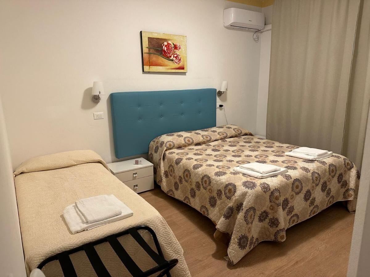A small, clean room with two beds, a blue headboard, a painting, and a white air conditioner.