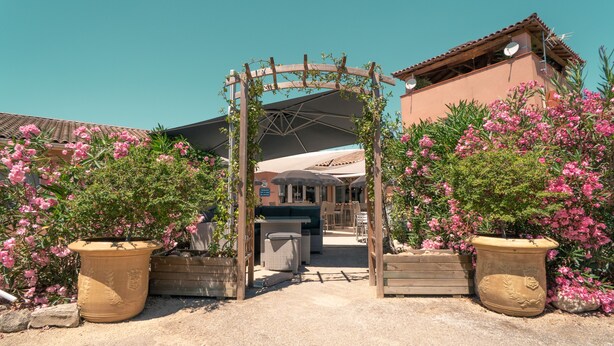 Un área de estar al aire libre con entrada cubierta por una pérgola, rodeada de plantas y flores en macetas.