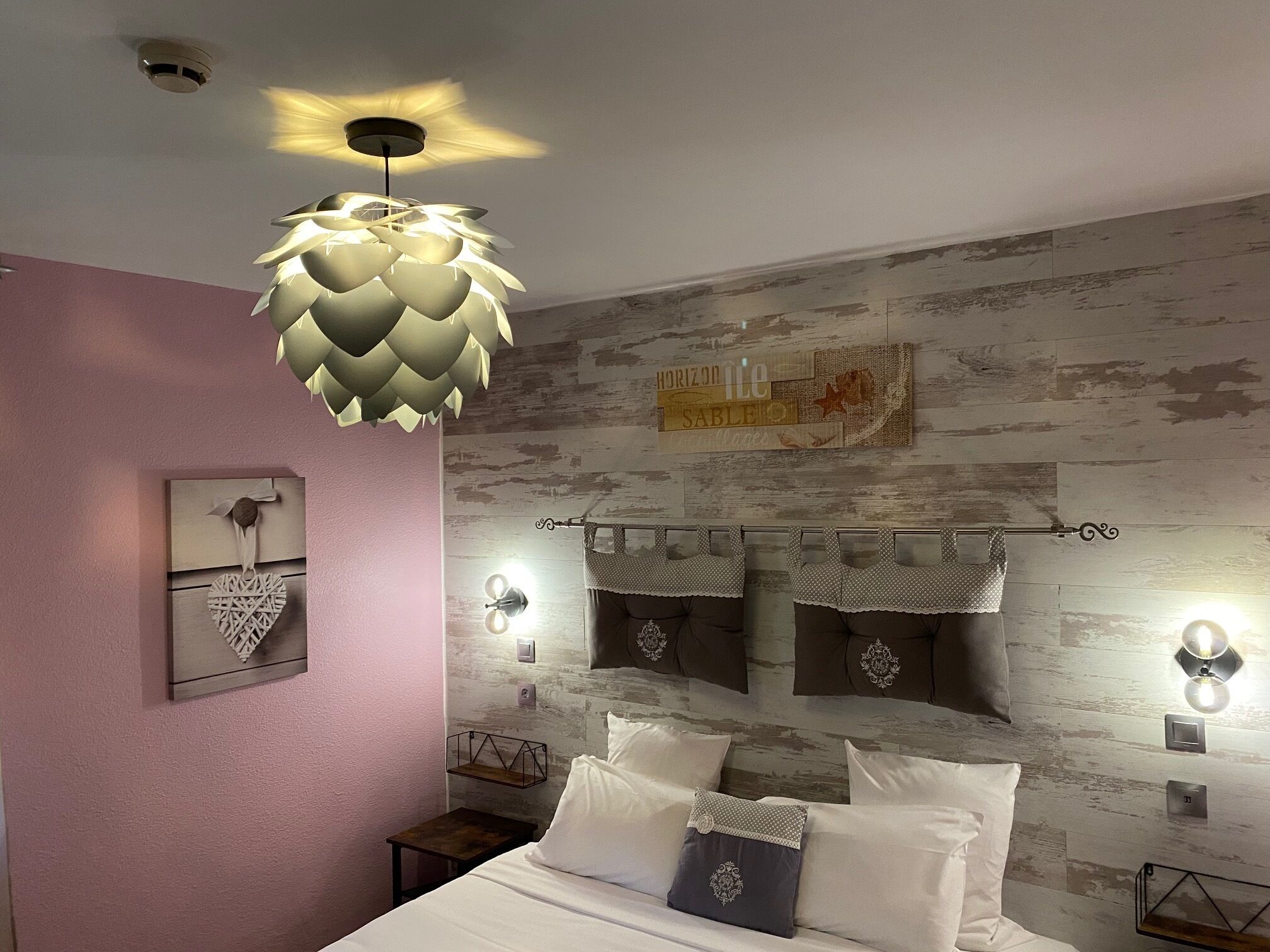 A bedroom with a bed, decorative pillows, a unique ceiling light, and a rustic wall design.