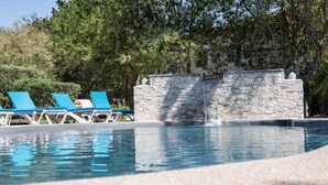 Zona de piscina con sillas reclinables azules, un muro de piedra y árboles al fondo.