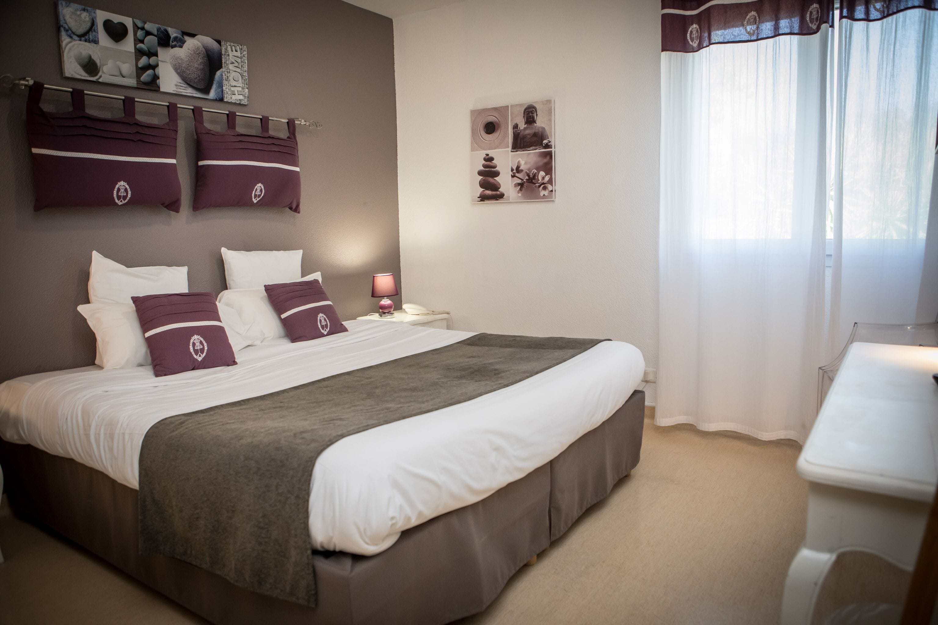 A neatly made bed with a grey bed skirt and white linens, a bedside table with a lamp, and a wall with framed artwork and a bulletin board.