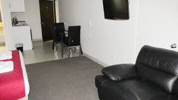A compact room with a black leather sofa, a small kitchenette, a dining area with chairs, and a flat-screen TV mounted on the wall.