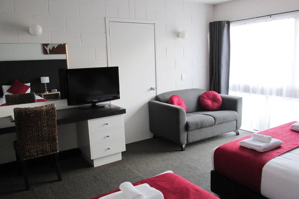 Et hotelværelse med seng, skrivebord med computer, stol, fjernsyn og et vindue med gardiner.