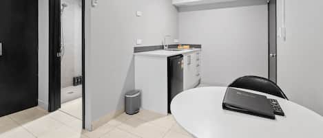 Una habitación de hotel moderna con una mesa blanca, una silla negra, una pequeña cocina y una ducha.