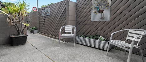 Un espacio exterior pavimentado con dos sillas de metal, una planta en maceta y una pared decorativa con un diseño geométrico.