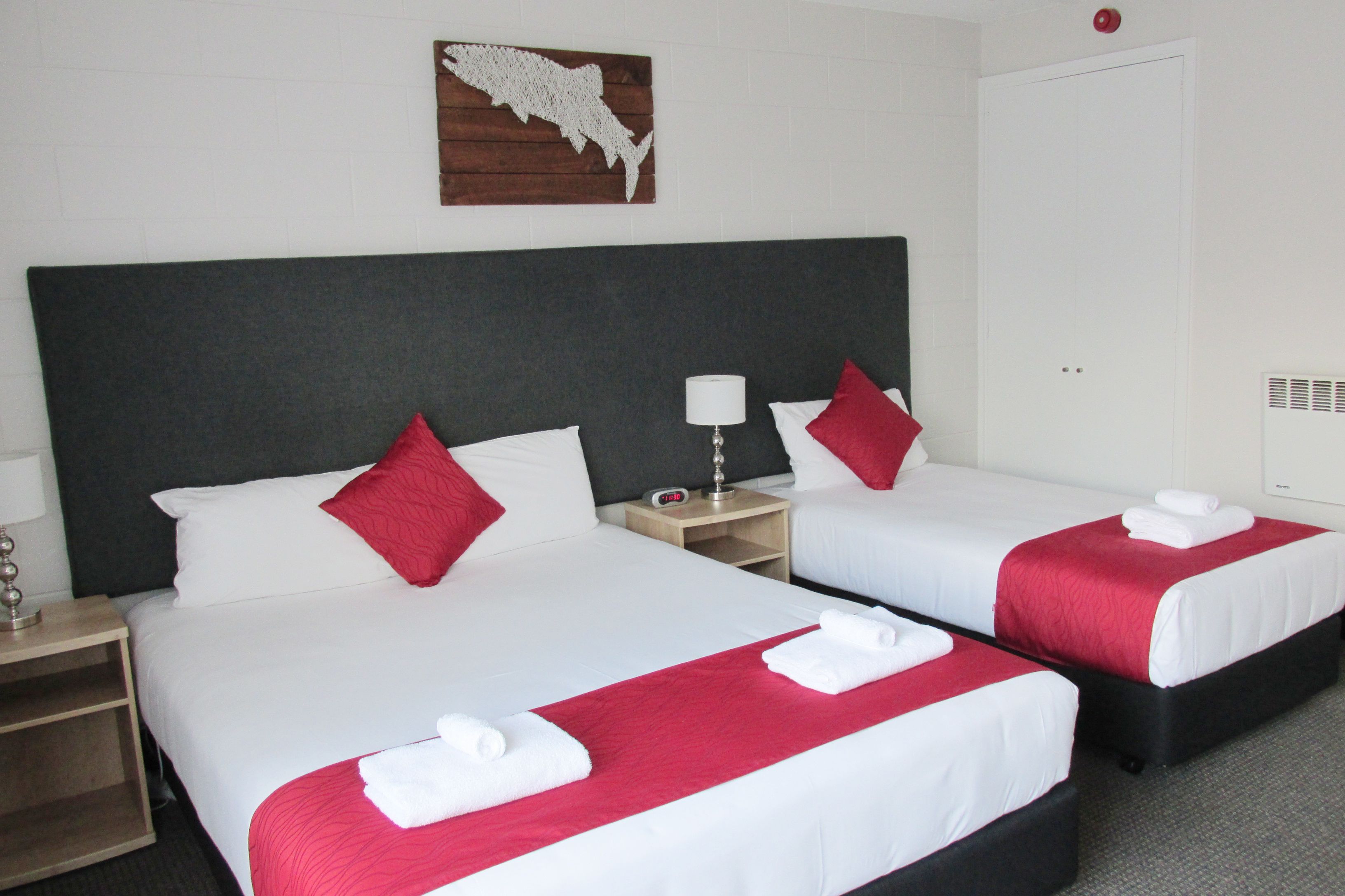 Hotelkamer met twee bedden, elk voorzien van witte beddengoed en rode accenten, een houten hoofdeinde, een nachtkastje met een lamp en een kunstwerk aan de muur.