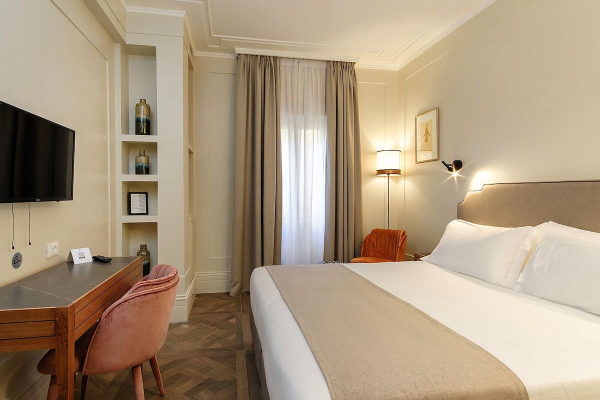 Une chambre d’hôtel comprenant un lit, un bureau avec une chaise, une télévision, une étagère avec des objets décoratifs, une lampe et un tableau encadré au mur.