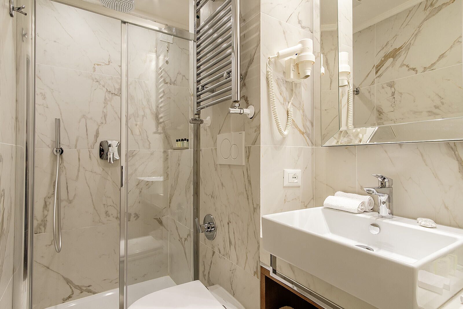 A modern bathroom with a glass-enclosed shower, a marble-tiled wall, a white sink, and a hairdryer mounted on the wall.