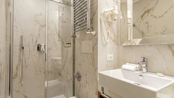 A modern bathroom with a glass-enclosed shower, a marble-tiled wall, a white sink, and a hairdryer mounted on the wall.