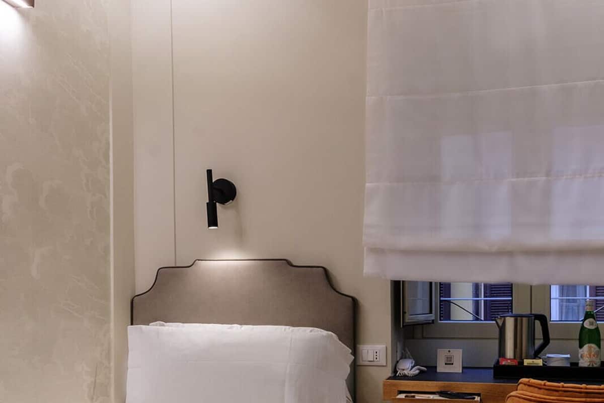 Une chambre d’hôtel de taille réduite, comprenant un lit simple, un petit bureau avec une chaise, une fenêtre avec des stores et un luminaire fixé au mur.