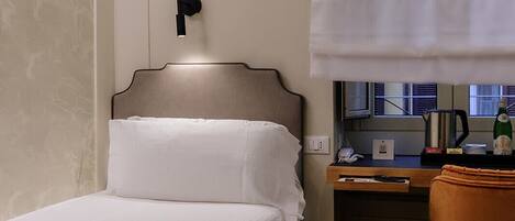 A compact hotel room with a single bed, a small desk with a chair, a window with blinds, and a wall-mounted light fixture.