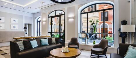 A modern hotel lobby with a checkered floor, a black leather sofa, a round wooden table with candles, and large arched windows offering a view of the street.