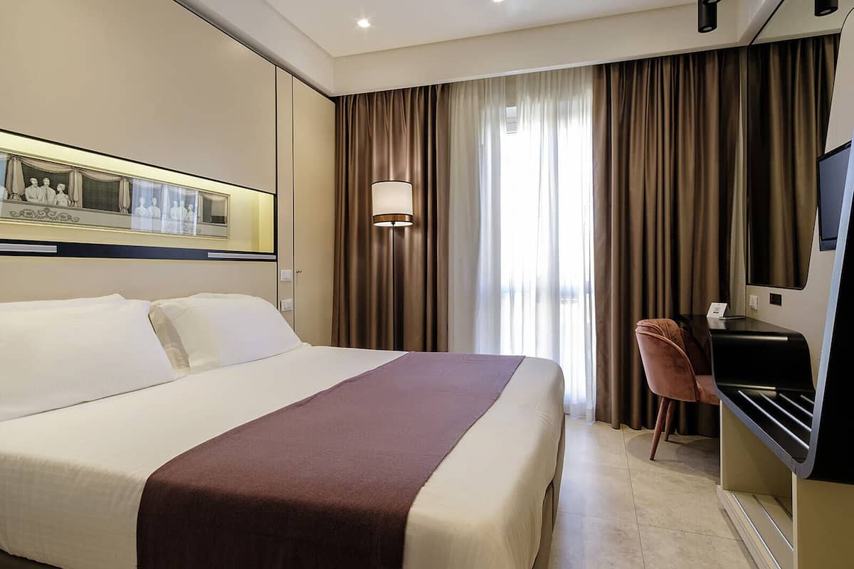 Une chambre d’hôtel avec un grand lit, un bureau avec une chaise, une télévision et une fenêtre avec des rideaux.
