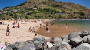A beach with people sunbathing and swimming, surrounded by large rocks and a hill with houses in the background. - Hotel White Waters (Machico)