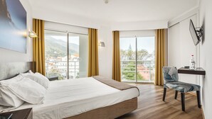 A modern hotel room with a large bed, a desk with a chair, and a view of the city through large windows. - Hotel White Waters (Machico)