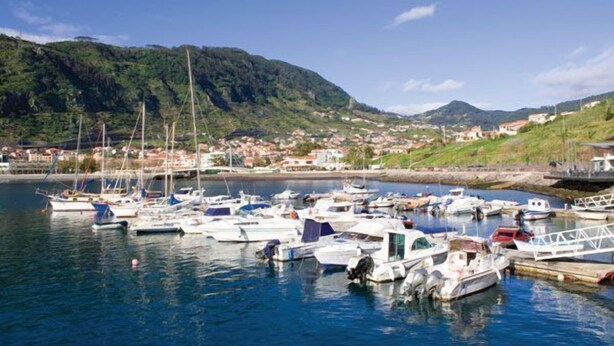A marina with numerous boats docked, surrounded by a hilly landscape and a small town in the background. - Hotel White Waters (Machico)