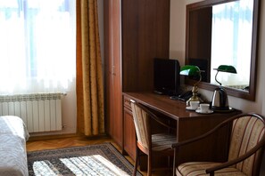 A hotel room with a wooden desk, a chair, a bed, a radiator, a window with curtains, and a mirror.