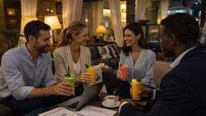 A group of people sitting around a table with laptops and documents, holding drinks, in a warmly lit indoor setting with a couch and curtains in the background. - Hotel Life Gran Via (Madrid)